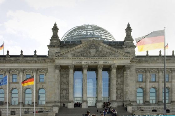 Aufnahme des Reichstagsgebäudes mit Glaskuppel in Berlin. Auf und vor dem Gebäude stehen mehrere Deuschlandflaggen und eine Europaflagge.