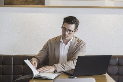 Ein junger Mann arbeitet am Laptop und liest ein Buch.
